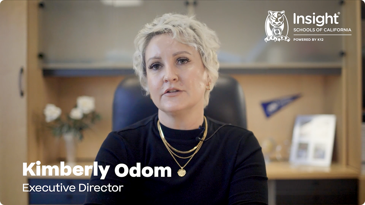 A person with short blonde hair, wearing a black top and gold necklaces, sits in an office. Text reads: "Kimberly Odom, Executive Director." The office background is blurred.